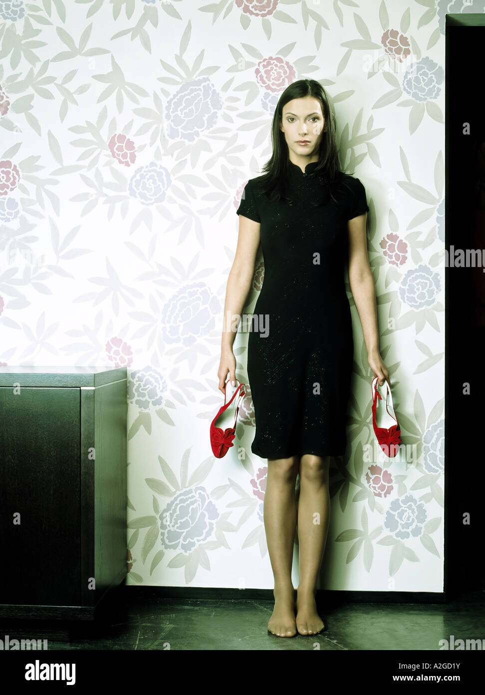 Indoor camera piatta giovane donna 25 30 brunette capelli lunghi vestito nero rosso scarpe scarpe tenere stand piedi piedi nudi Foto Stock