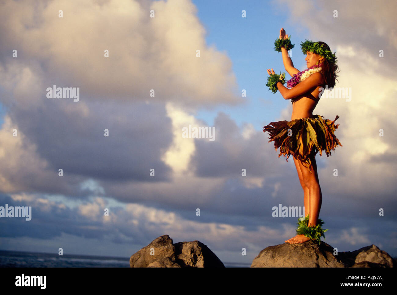 Hawaiani nativi donna che indossa un mantello di erba foglia di felce bracciali e motivi floreali colorati leis sorge su una roccia che si affaccia sull'oceano Foto Stock