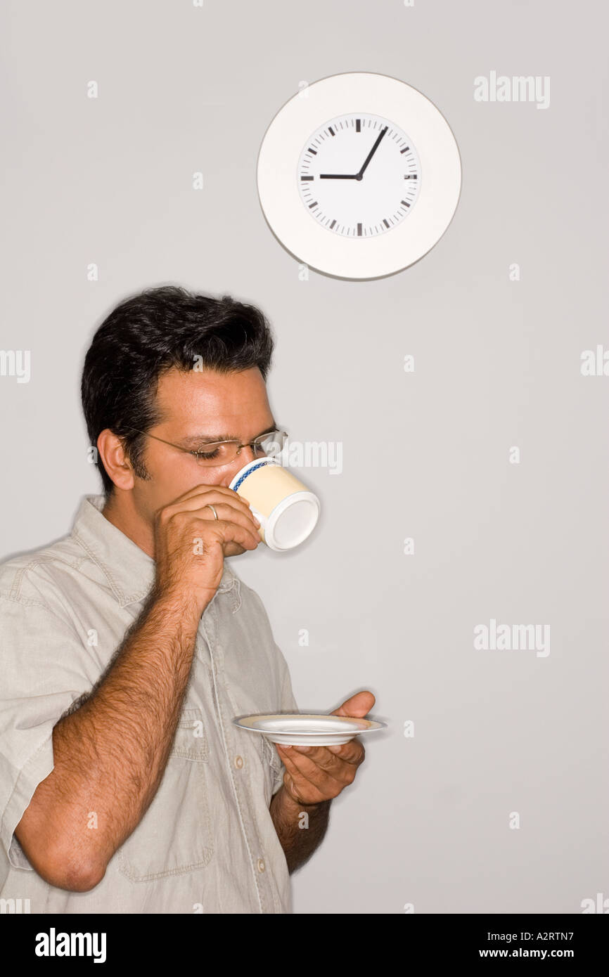 Uno dai capelli scuri giovane uomo è in piedi di fronte a un muro di bere il caffè o il tè. L'orologio è di cinque (5) minuti passato Foto Stock
