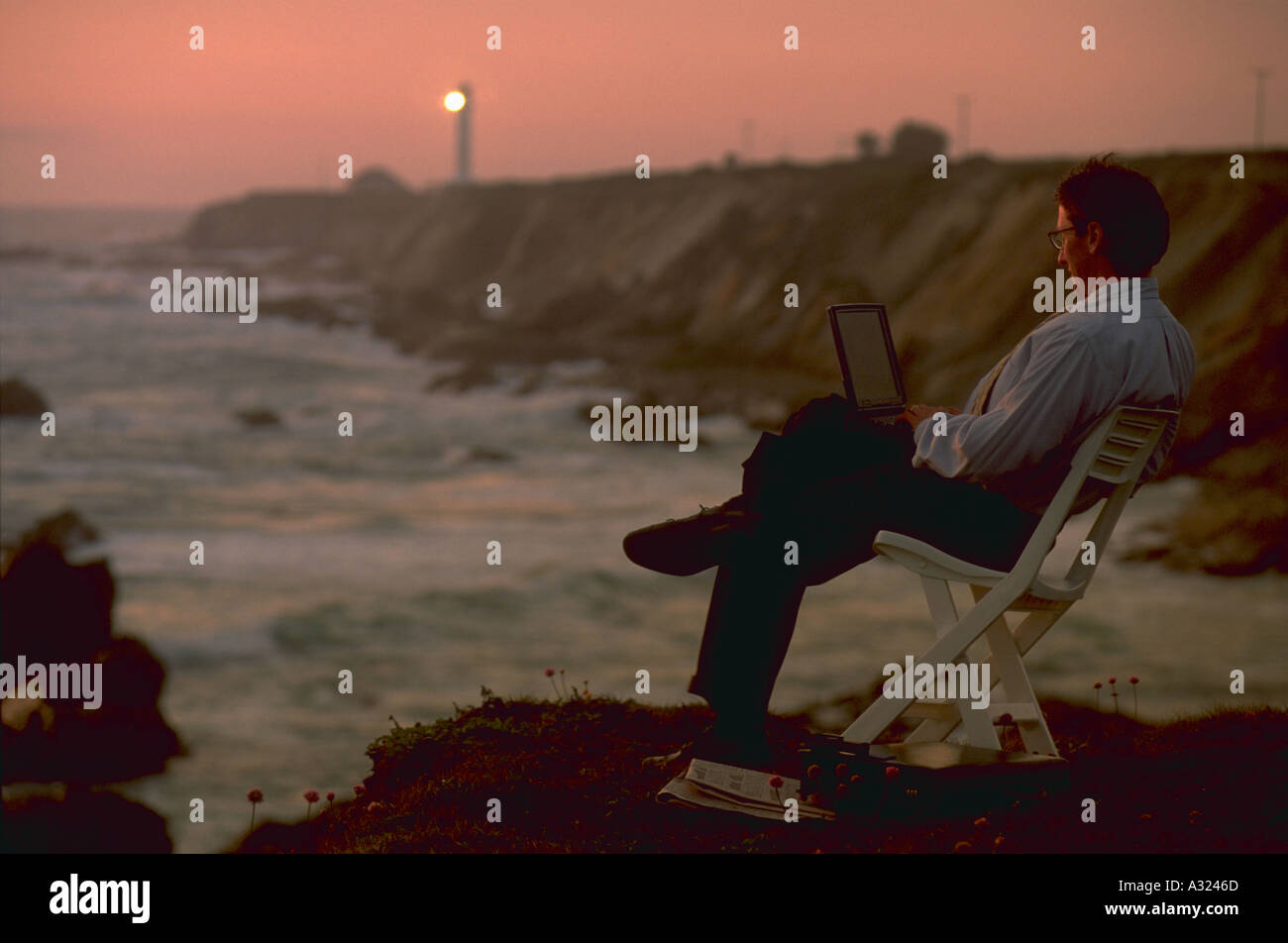 Uomo d affari con computer portatile e seduto nella sedia sulla costa rocciosa del punto Arena California con faro dietro Foto Stock