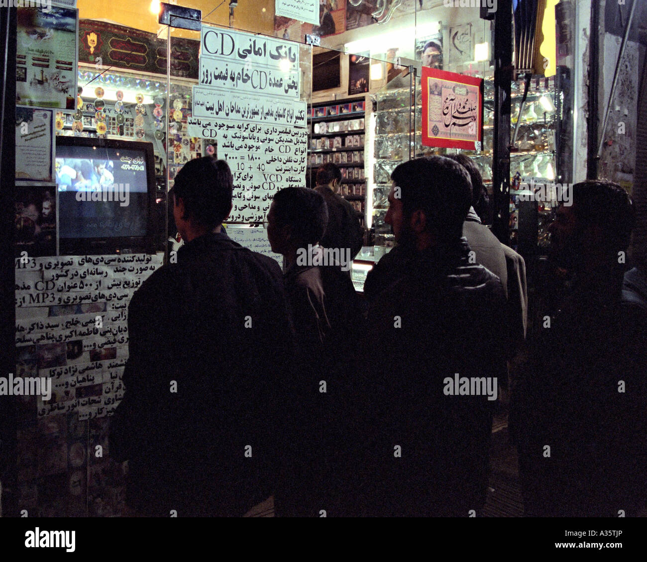 Un gruppo di uomini e ragazzi guarda un programma religioso al di fuori della finestra di un negozio che vende i religiosi nastri e CD. Qom, Iran Foto Stock