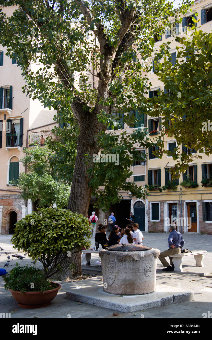 Italia Veneto Venezia Novo Ghetto Ebraico Ghetto Nuovo adulti e bambini seduti sotto un albero accanto a un tappate bene acqua in piazza Foto Stock