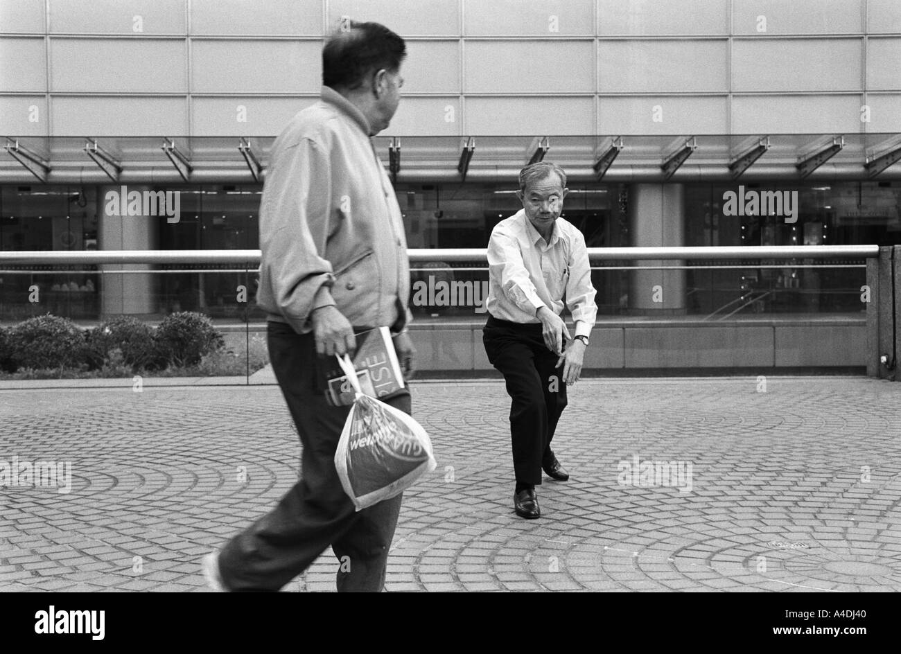 Un uomo con una borsa di trasporto passa un uomo che sta facendo il suo Tai Chi al di fuori del suo ufficio edificio prima del lavoro di Hong Kong. Foto Stock