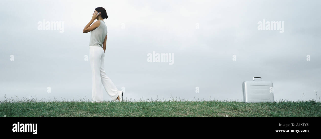 Donna che cammina su erba parlando al cellulare, valigetta metallica sul terreno dietro di lei Foto Stock