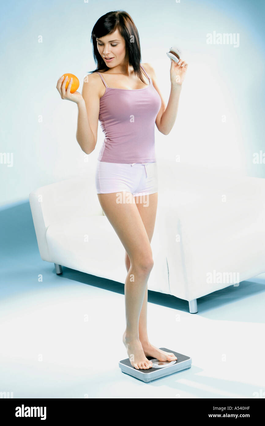 Giovane donna in piedi su una bilancia con il biscotto di arancione in mano Foto Stock