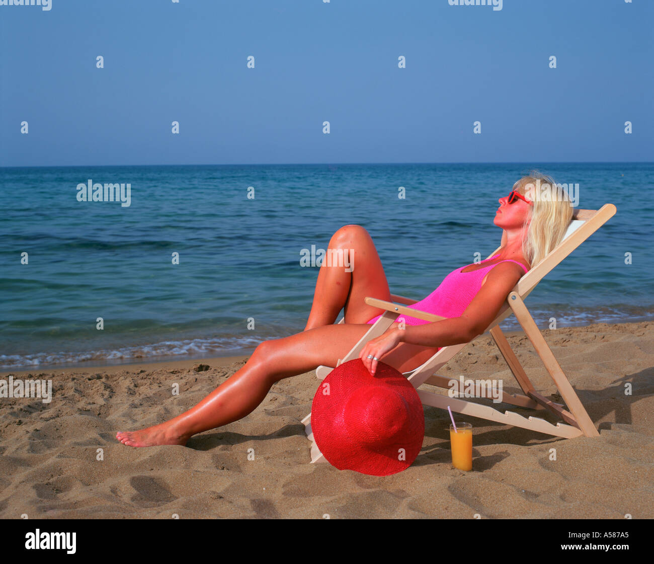 Donna bionda rosa in costume da bagno seduto su una sedia a sdraio sulla spiaggia e tenendo un grande Sun Red Hat Foto Stock