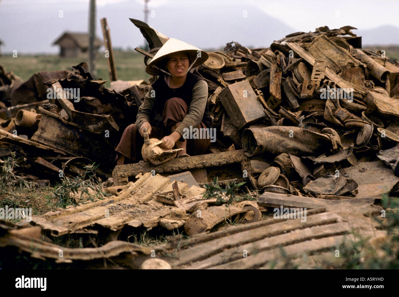 Il Vietnam, una donna di manodopera con cappello conico smantella rottami di metallo con un martello che lavora davanti di cumuli di Rusty materiale bellico Foto Stock