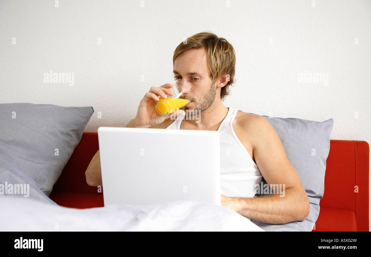 Giovane uomo utilizzando laptop su letto di bere succo di frutta Foto Stock