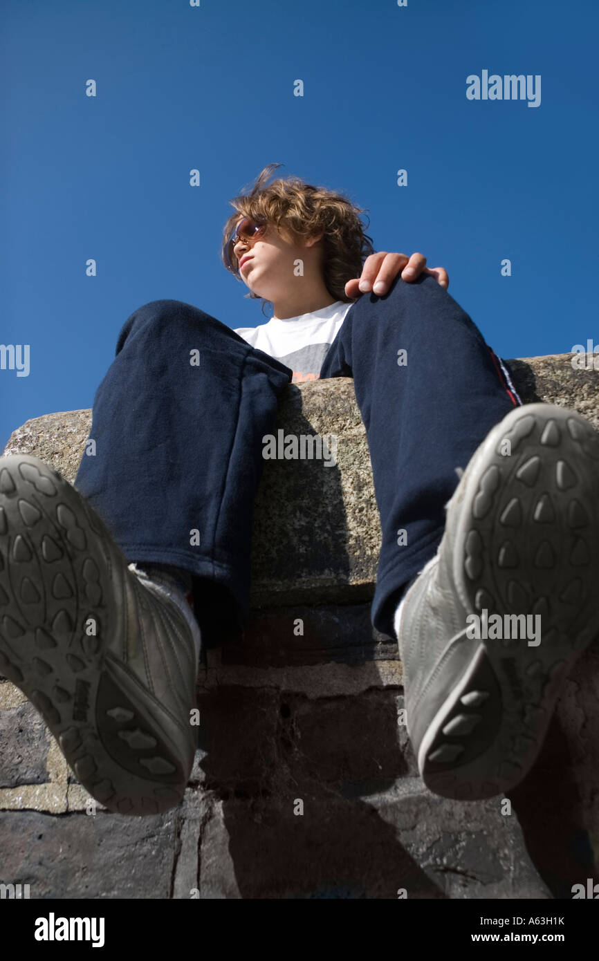 Guardando verso l'alto un ragazzo adolescente seduto su un muro di pietra il bambino ha ha i capelli lunghi e indossa occhiali scuri Foto Stock