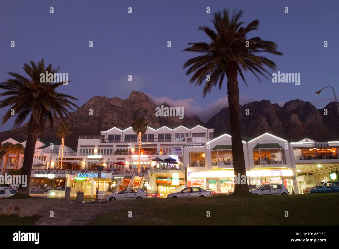 La trafficata strada principale di Camps Bay Città del Capo Sud Africa che mostra i ristoranti e i negozi che si trovano di fronte all'acqua Foto Stock