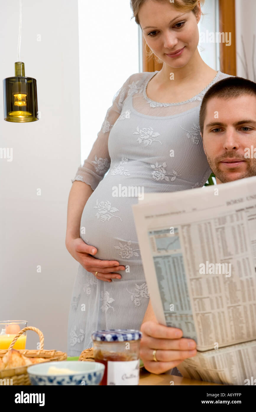 Donna incinta in piedi accanto ad un uomo che legge un giornale a colazione Foto Stock