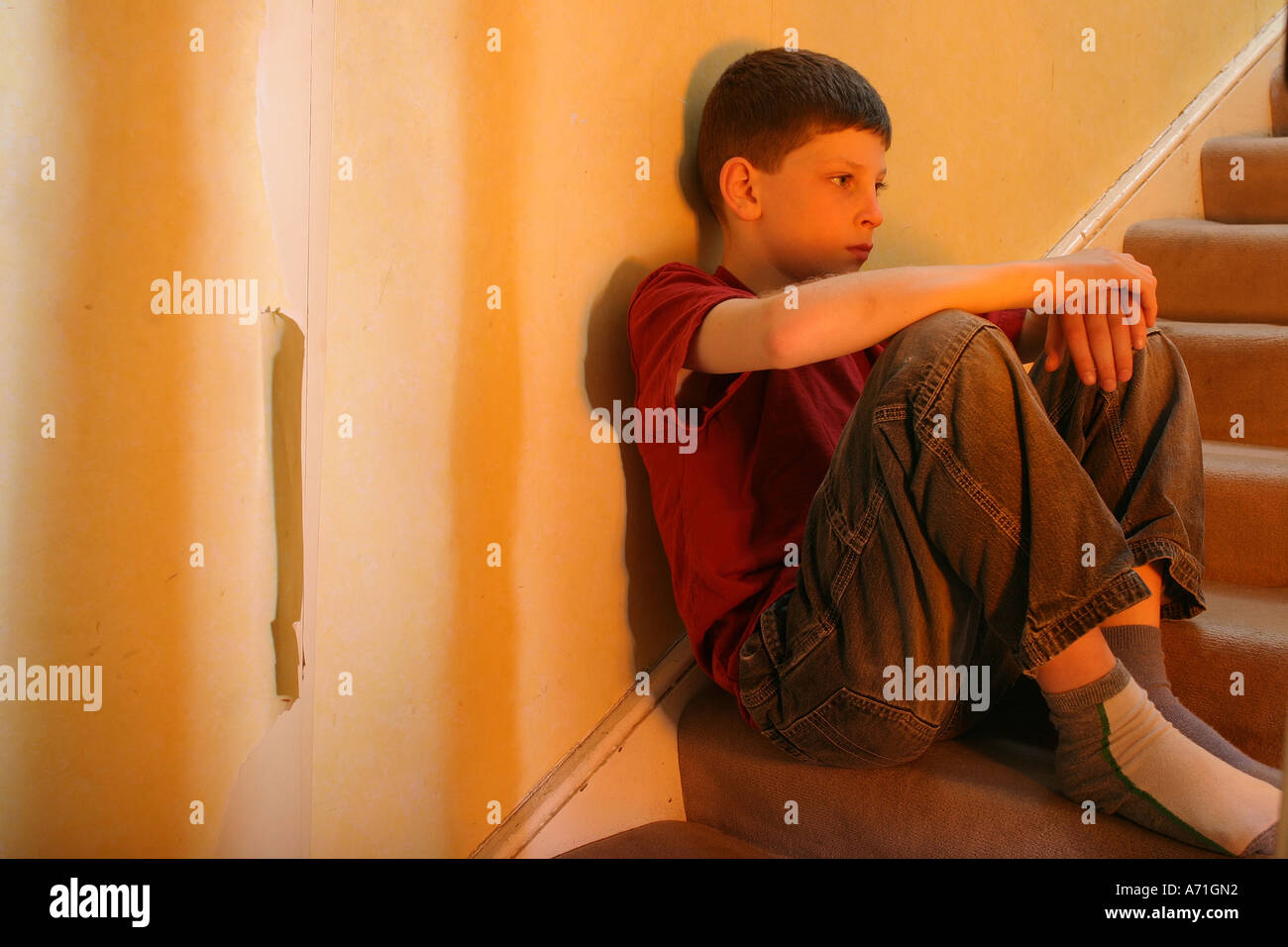 Un riflettente e rattristato 8 anno vecchio ragazzo seduto sulle scale di casa con parete strappati carta in background. Foto Stock