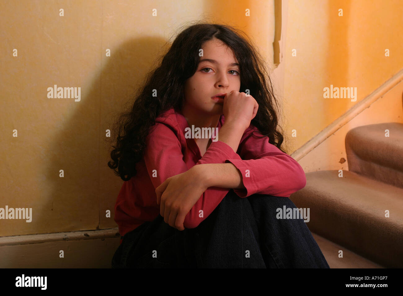 Un riflettente e rattristato 10 enne ragazza seduta da scale a casa con parete strappati carta in background. Foto Stock