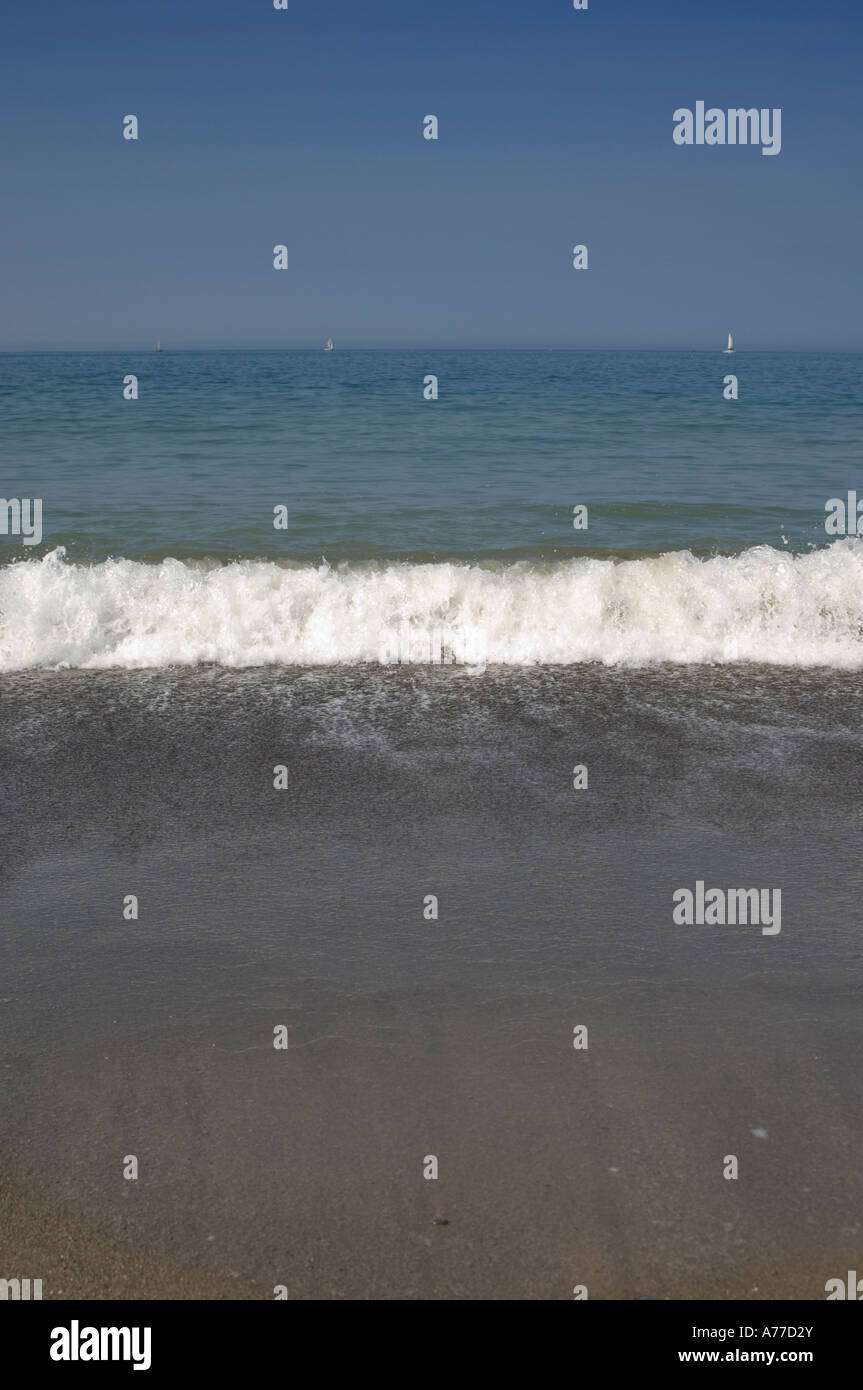 Piccole onde che si infrangono sulla spiaggia in una calda giornata estiva con cielo azzurro e yacht all'orizzonte del Galles cymru Foto Stock