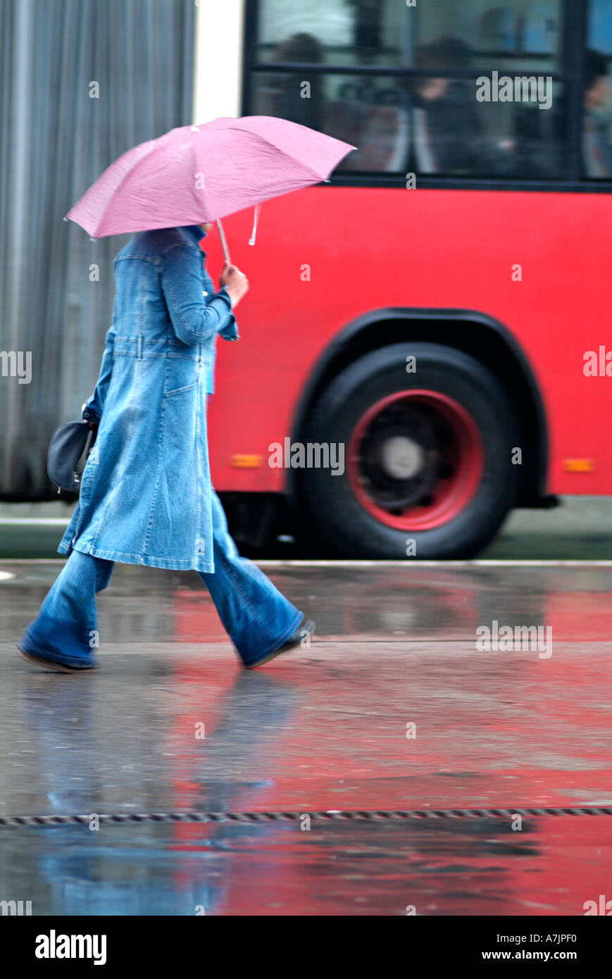 Donna che cammina lungo un marciapiede della città di fronte ad un bus rosso durante una doccia a pioggia Foto Stock
