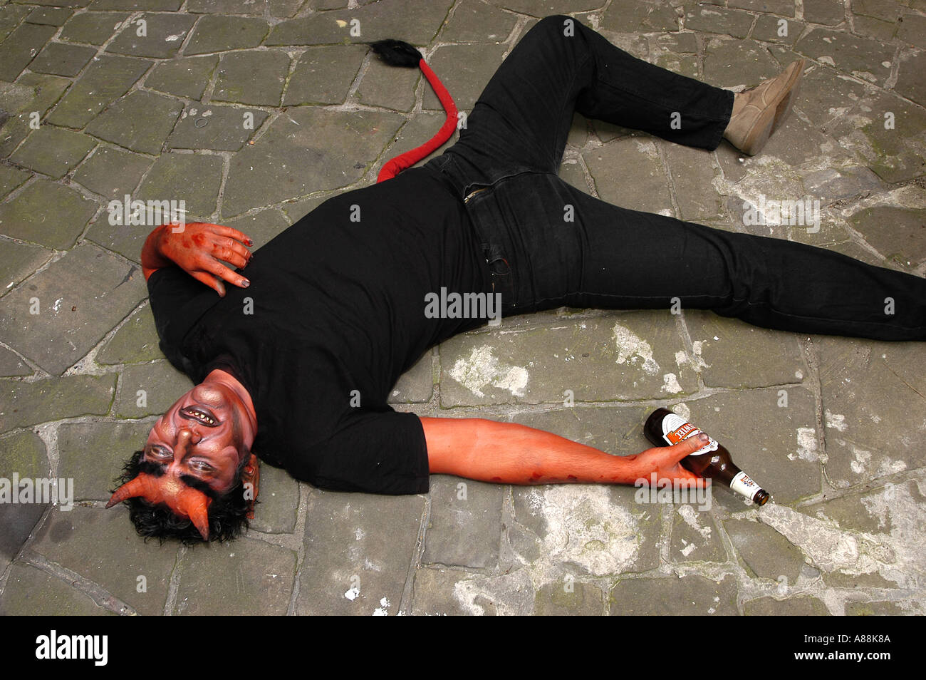 Il diavolo maschile in costume giace su un marciapiede di pietra con una bottiglia di birra vuota, indossa abiti neri e una coda rossa. Foto Stock