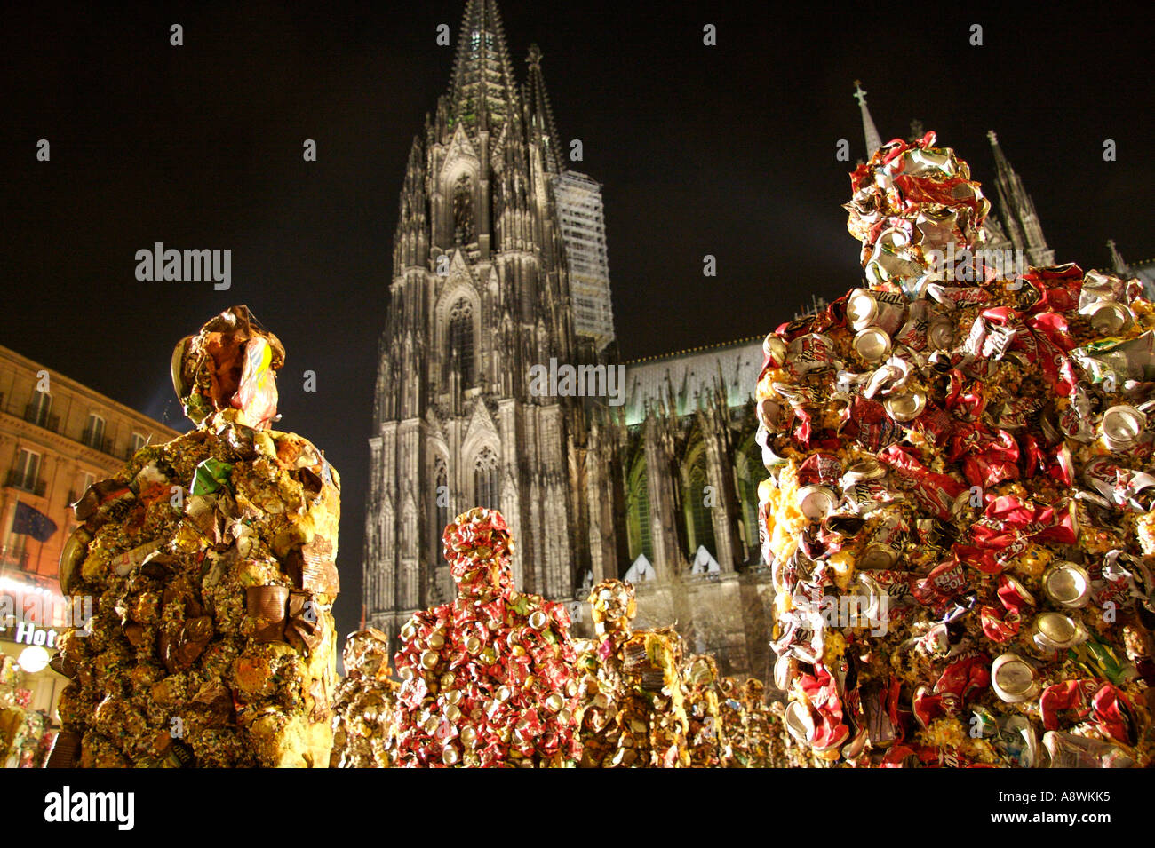 Di notte, figure dell’installazione ‘Trash People’ di HA Schult si trovano davanti alla cattedrale di Colonia illuminata. La figura in primo piano a destra è interamente rivestita di lattine di Coca-Cola rosse accartocciate, mentre le figure a sinistra sono realizzate con scarti di lamiera nei toni del marrone ruggine e dell’oro. Colonia, Renania Settentrionale-Vestfalia, Germania Foto Stock