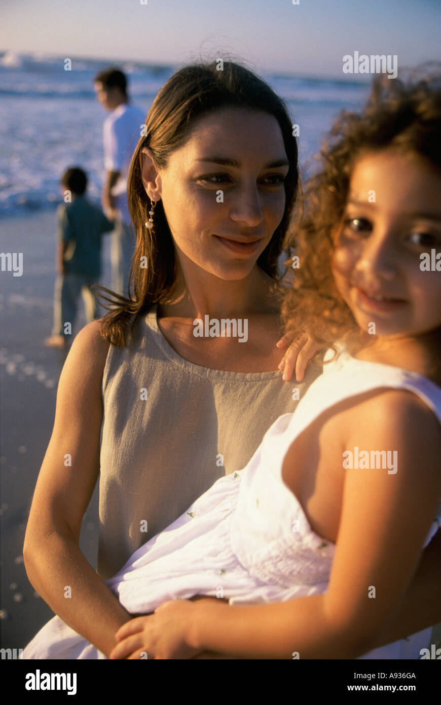 Ritratto di una madre tenendo la figlia sulla spiaggia con il padre e il figlio dietro di loro Foto Stock