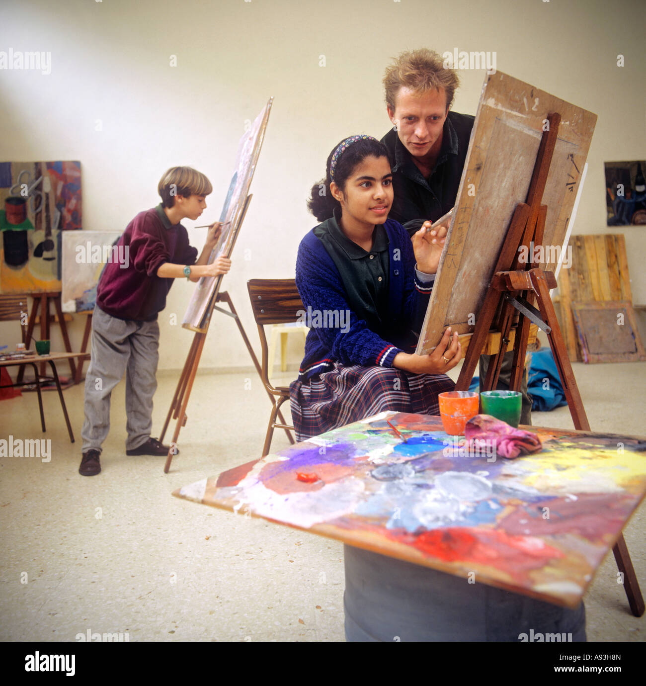 Arte classe Junior insegnante della scuola e junior girl allievo lavora su un dipinto ad acquerello in una scuola tecnica aula con ragazzo lavorando su una tela dietro Foto Stock