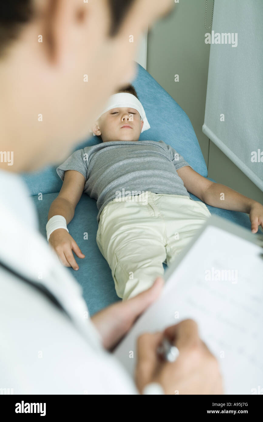 Ragazzo disteso sul lettino con bendaggio sulla fronte e il braccio, medico iscritto negli appunti Foto Stock