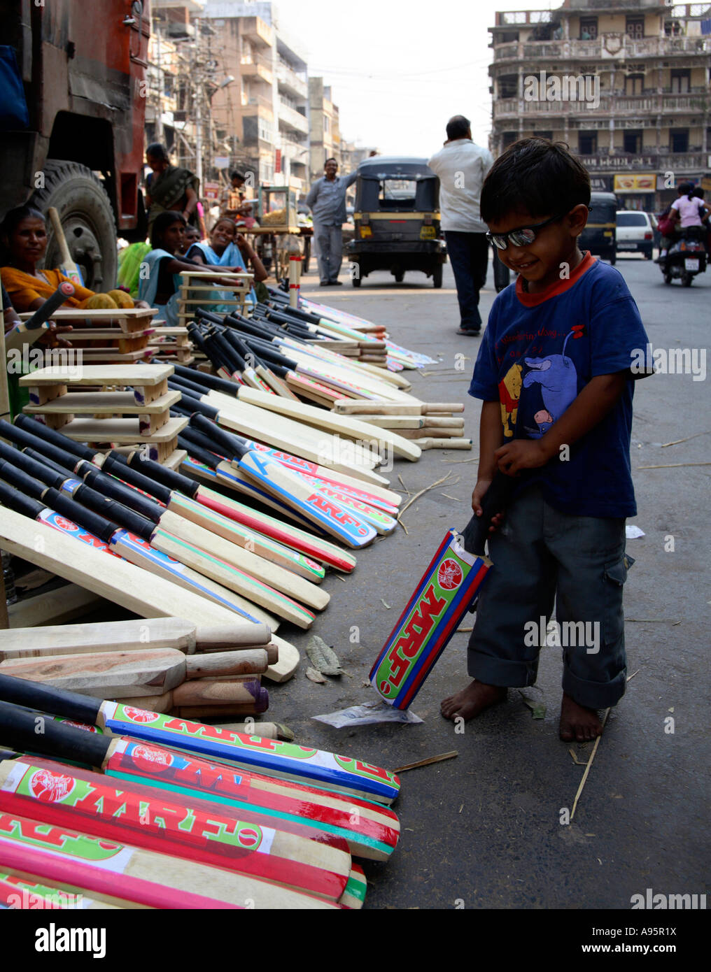 Giovane ragazzo in posa accanto alle donne la vendita di mazze da cricket a lato della strada, di Vadodara, Gujarat, India Foto Stock