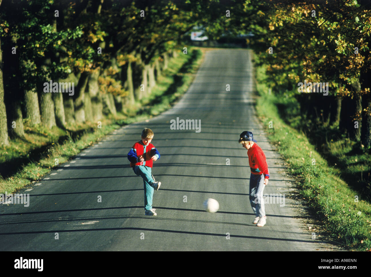 Ragazzi 7-11 anni in estate calci palla calcio (calcio) su un viale alberato strada di campagna Foto Stock