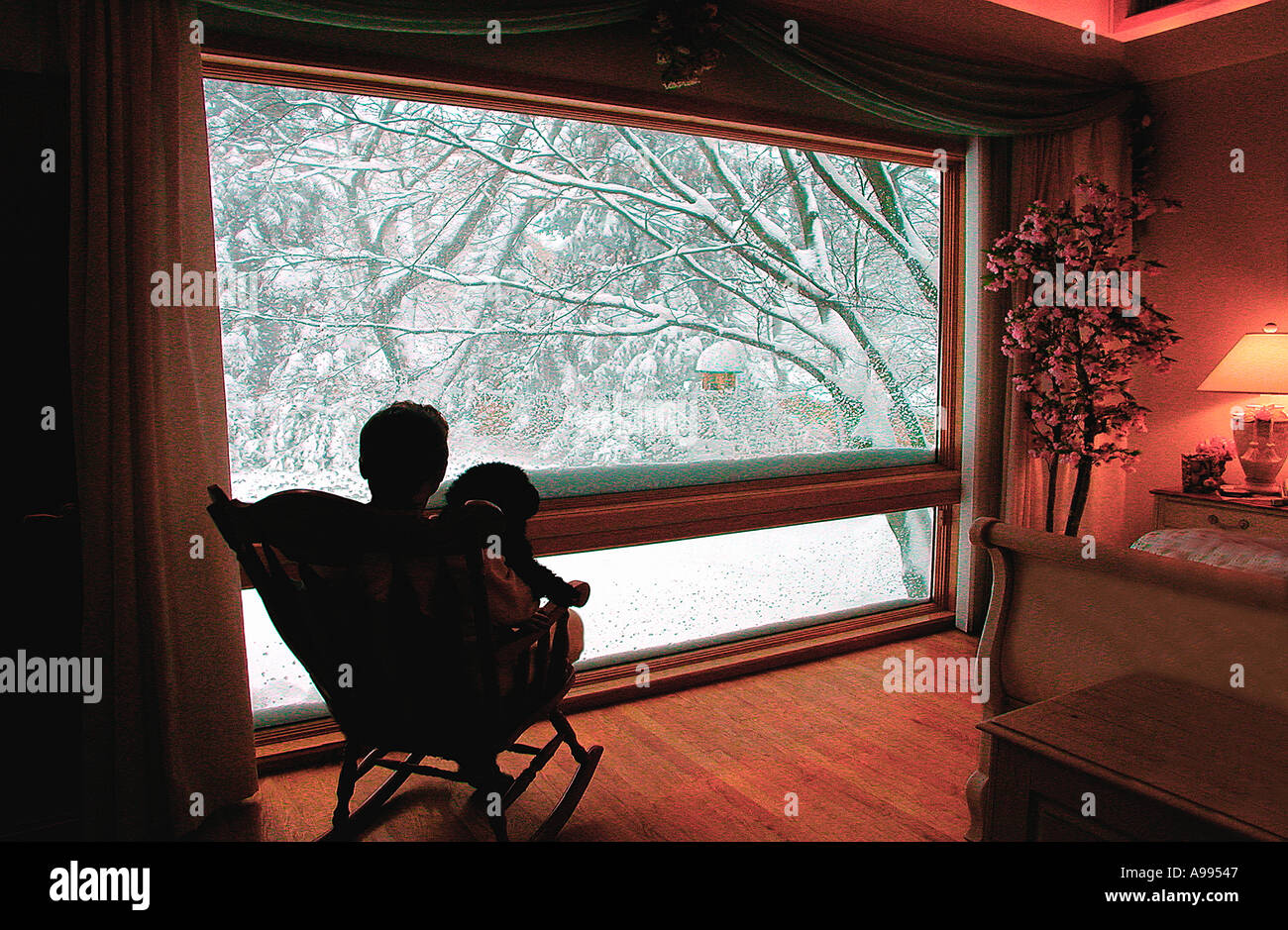 Un uomo seduto con il suo cane in una sedia a dondolo di fronte ad una grande finestra panoramica che guarda al coperto di neve alberi Foto Stock