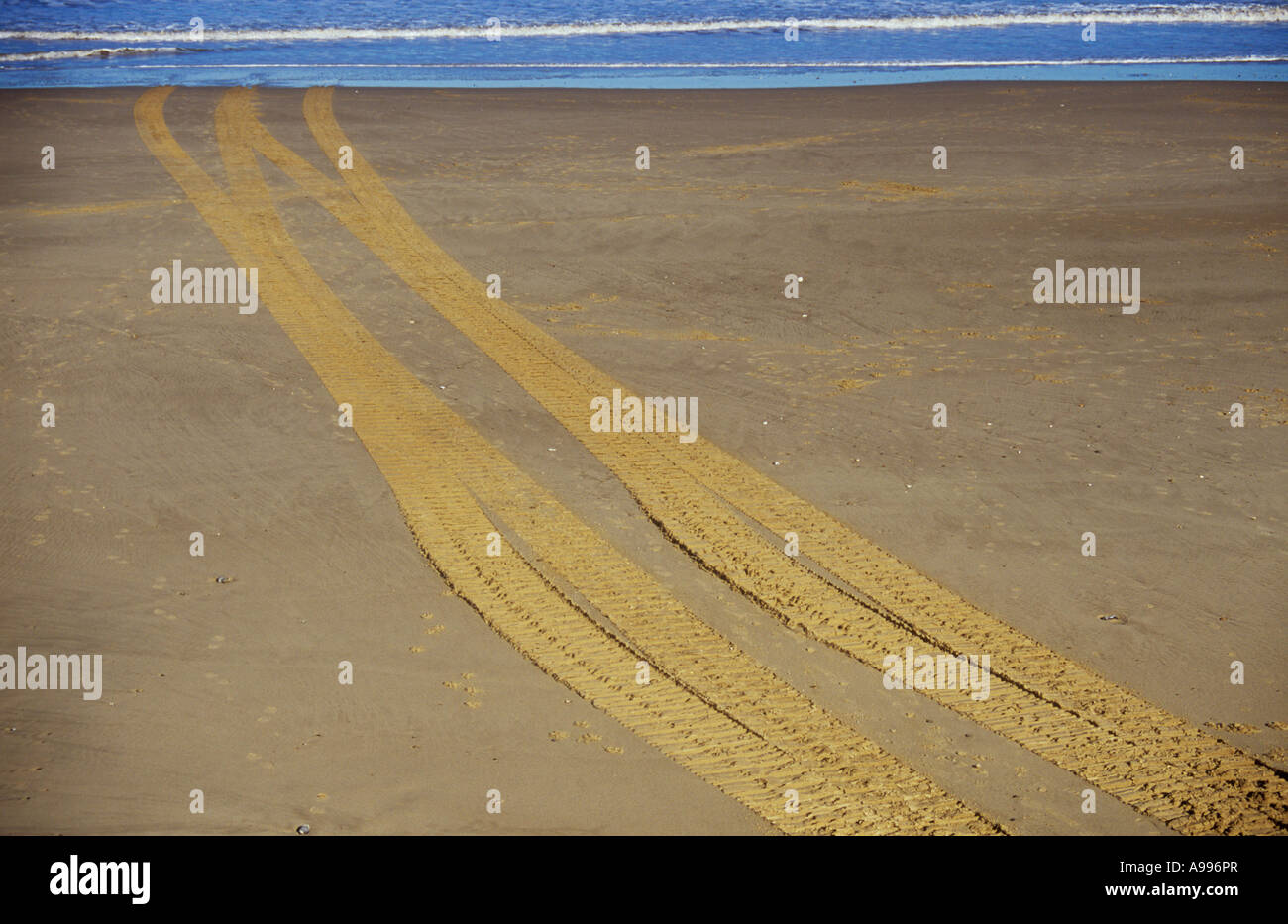 Un set di pneumatici o cingoli attraversando una chiara spiaggia sabbiosa e scomparire in o emergente da un delicato blu del mare Foto Stock