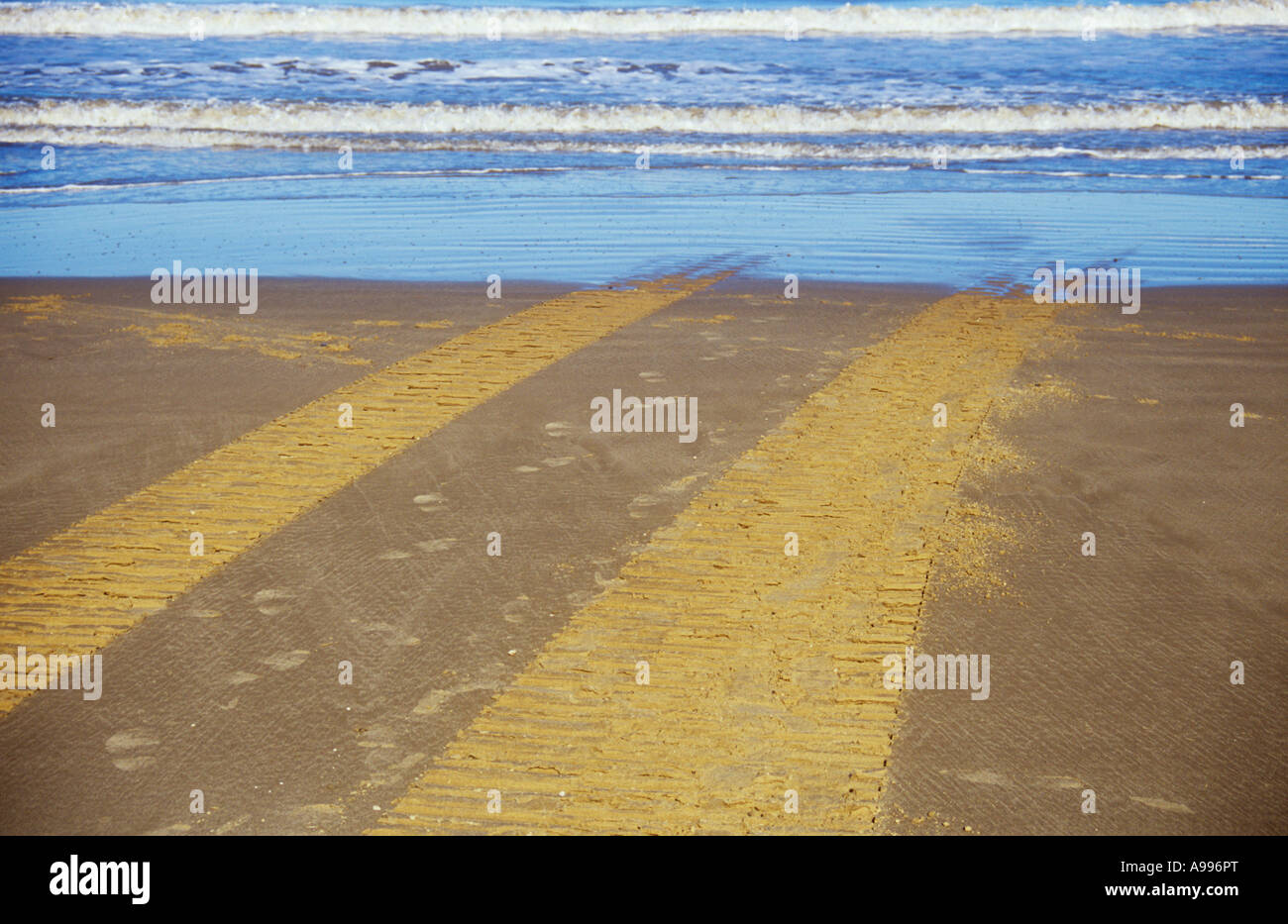 Un set di pneumatici o cingoli attraversando una chiara spiaggia sabbiosa e scomparire in o emergente da un delicato blu del mare Foto Stock