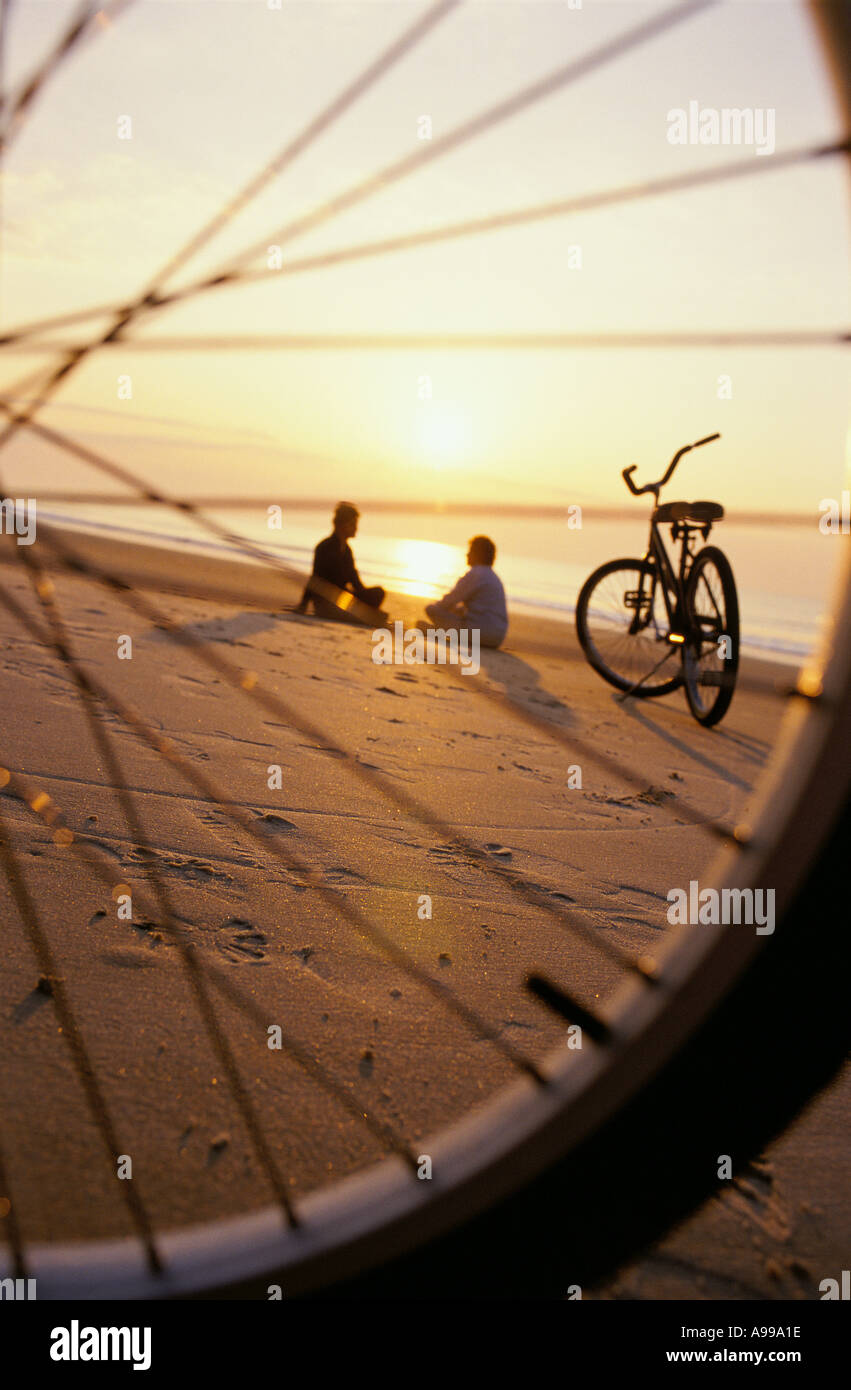 Sagome di due donne senior seduti insieme su una spiaggia all'alba come visto attraverso i raggi di una delle loro biciclette Foto Stock