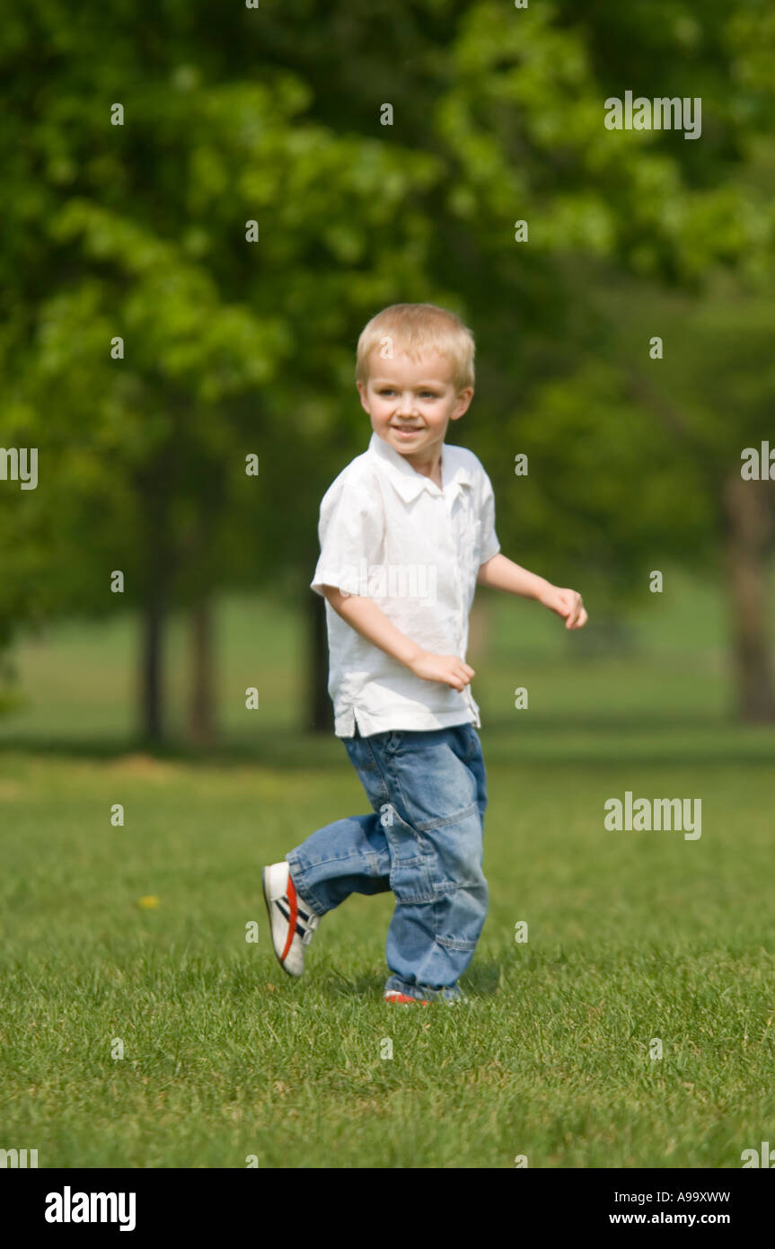 Un cheeky bionda e giovane ragazzo che indossa una camicia bianca e jeans, acceso e sorridente nel parco su una soleggiata giornata di primavera. Foto Stock