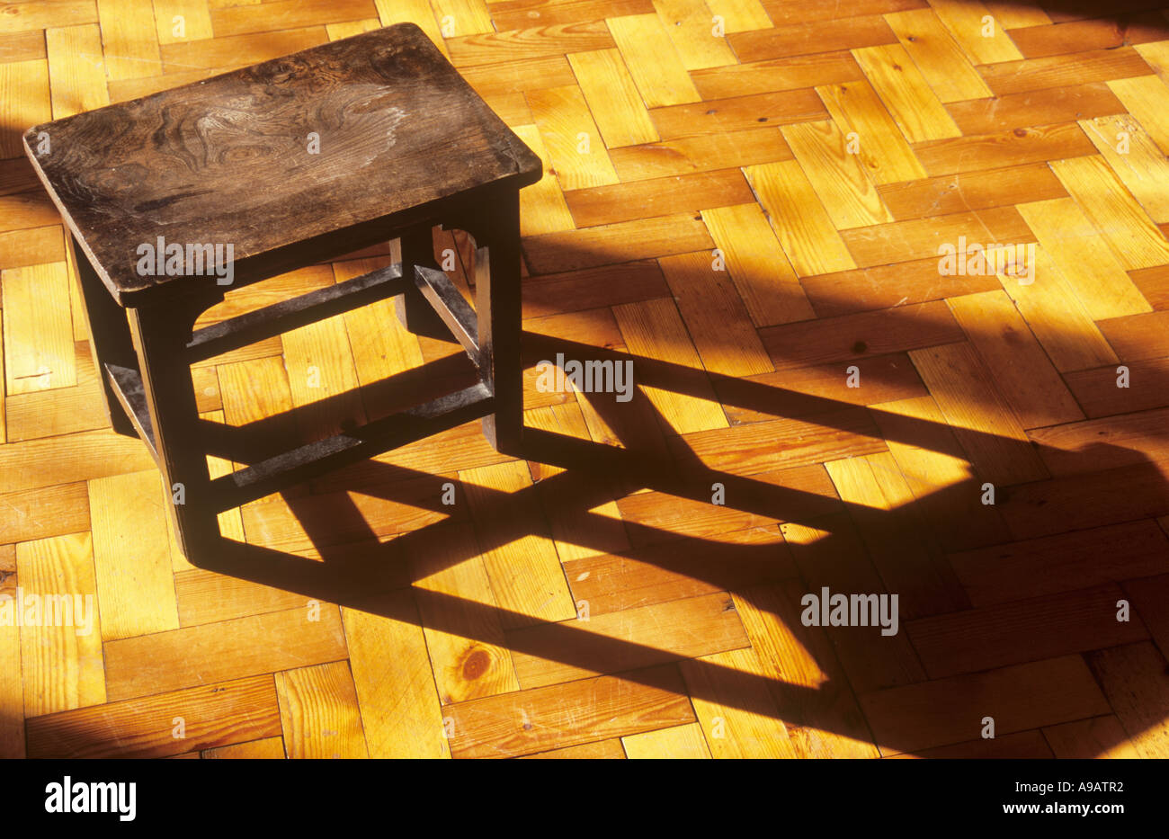 Squat Sgabello in legno o piccolo tavolo colata di una lunga ombra nella calda luce dorata attraverso un legno parquet pavimento di blocco Foto Stock