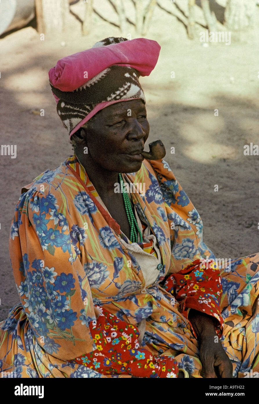 Herero donna che indossa gli abiti tradizionali e copricapo di fumare un tubo Foto Stock