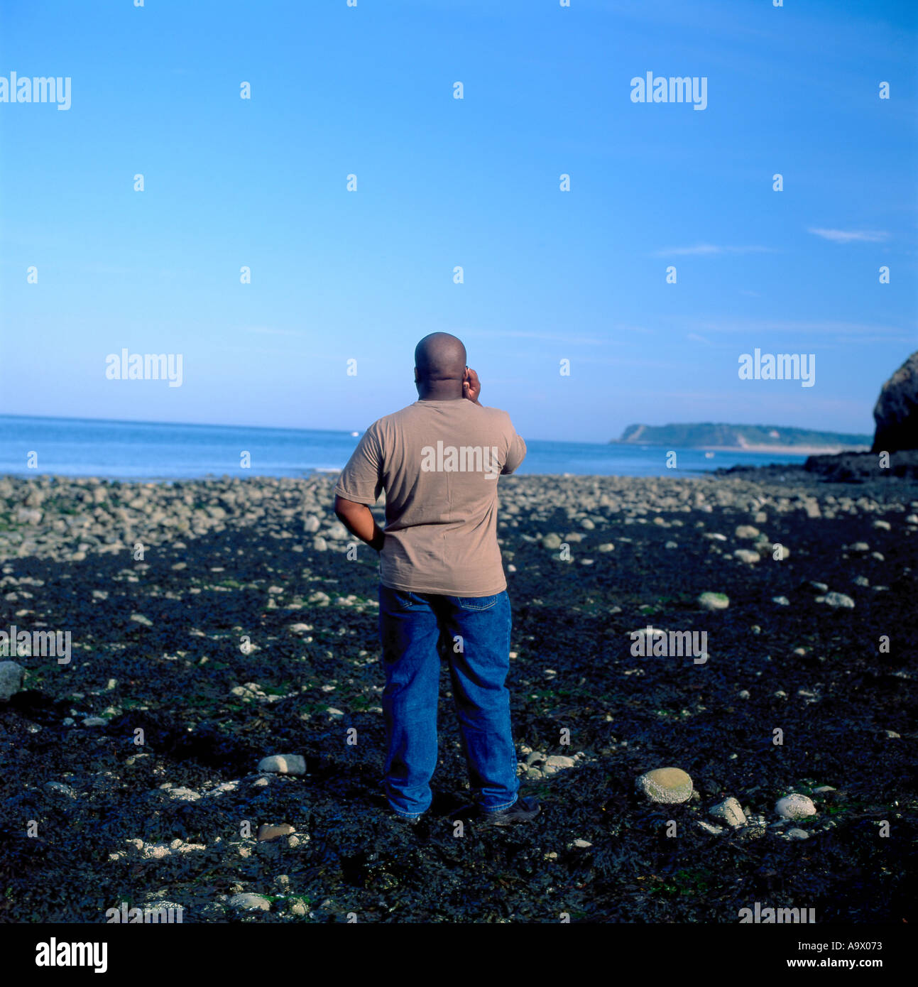Vista posteriore dell'uomo nero che parla sul telefono cellulare alla spiaggia e cielo blu copia spazio vicino Tenby Pembrokeshire Galles UK KATHY DEWITT Foto Stock