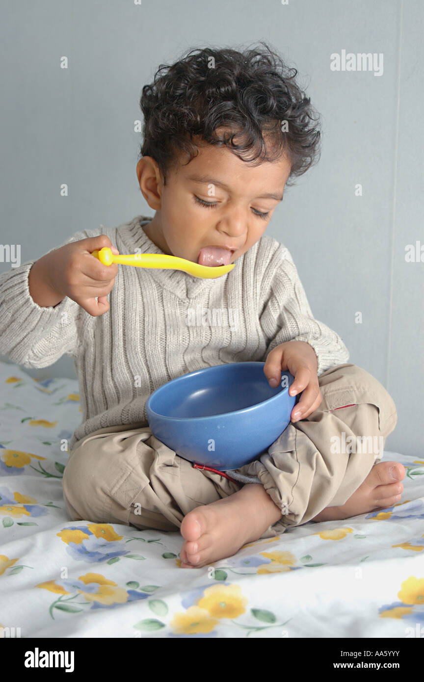 Ragazzo leccante, piccolo ragazzo indiano di due anni e mezzo che indossa un maglione bianco mangiando con cucchiaio giallo dalla ciotola blu Foto Stock