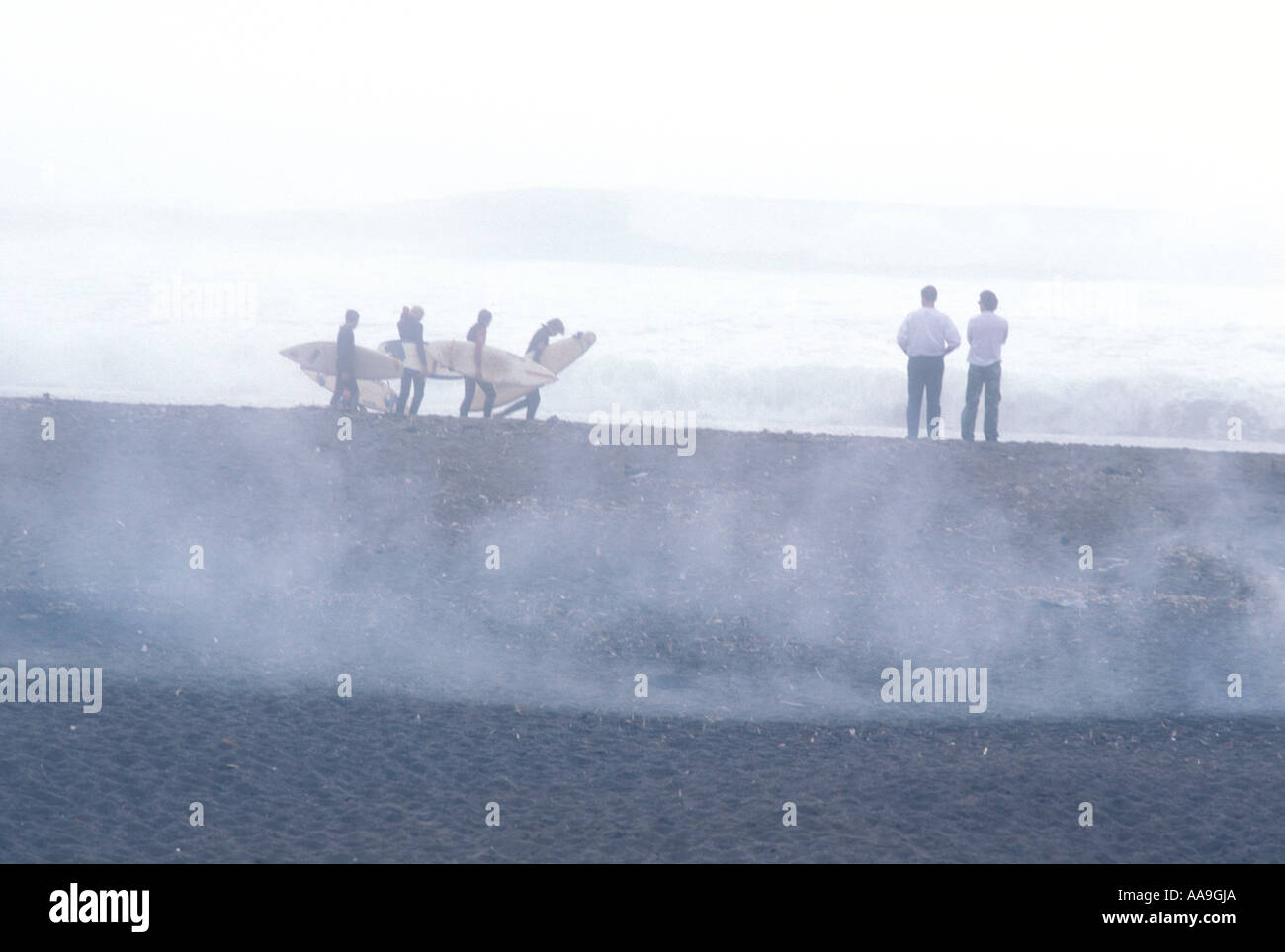 Surfers portano le loro schede su una spiaggia all'alba come nebbia sorge dalla sabbia e veli sulle colline di fronte alla baia. Foto Stock