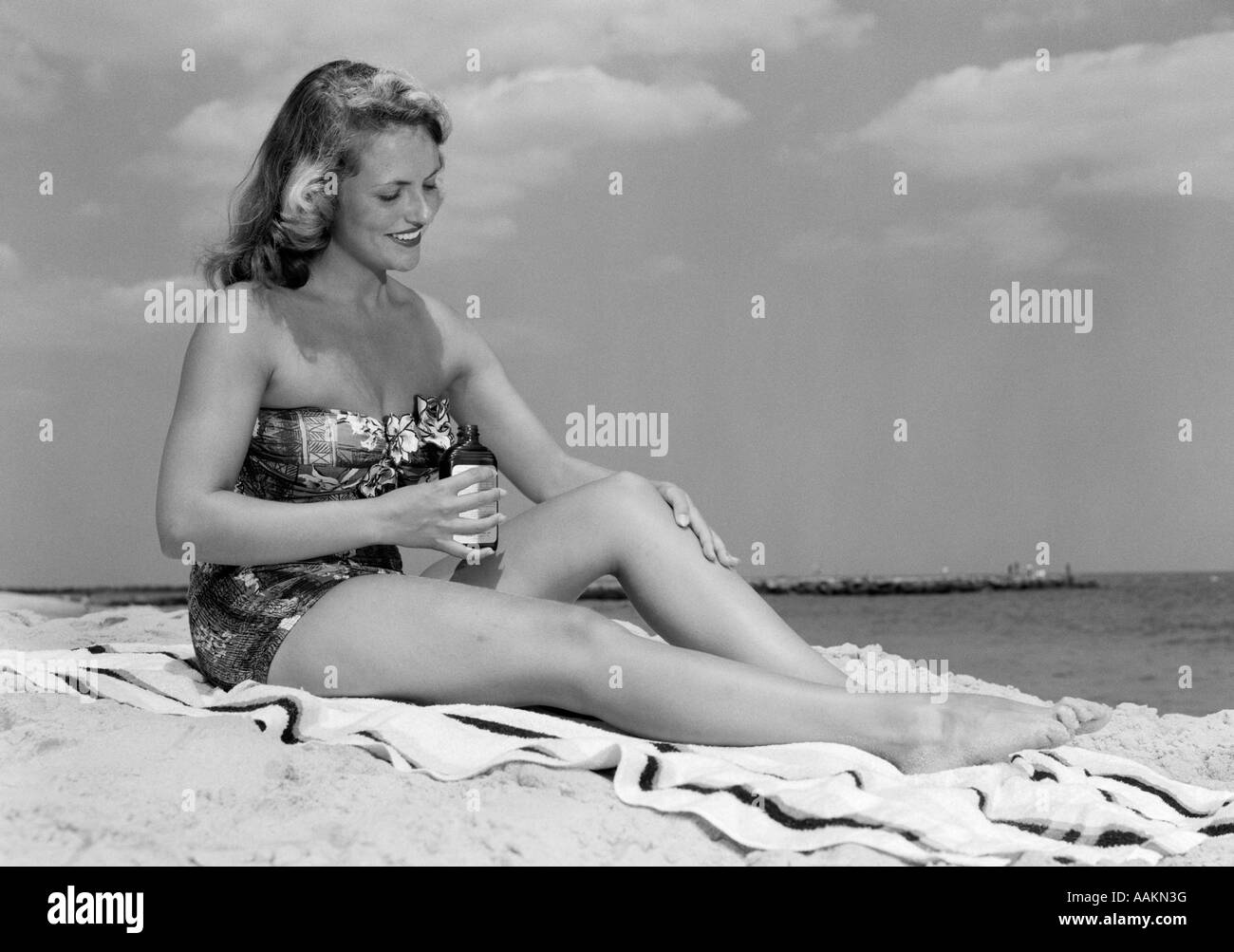 Anni Cinquanta la donna a senza spalline in pezzo unico COSTUME DA BAGNO seduto sul telo da spiaggia mettendo su una lozione solare Foto Stock