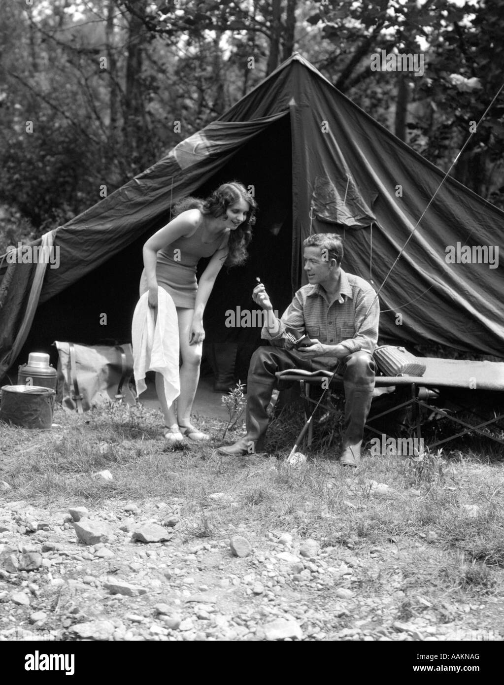 1920s CAMPING giovane uomo seduto dalla tenda con canna da pesca donna in piedi indossando COSTUME DA BAGNO Foto Stock