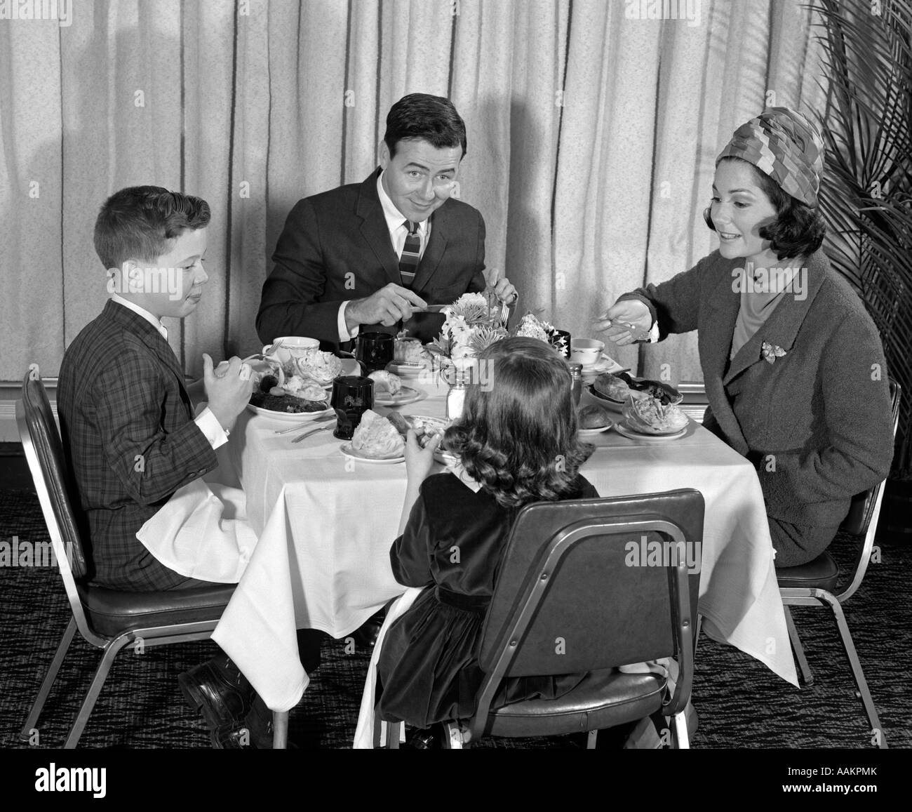 Anni sessanta sorridendo felice famiglia di quattro mangiare pasto nel ristorante mamma indossando turbante STYLE HAT rivolgendosi a sua figlia Foto Stock