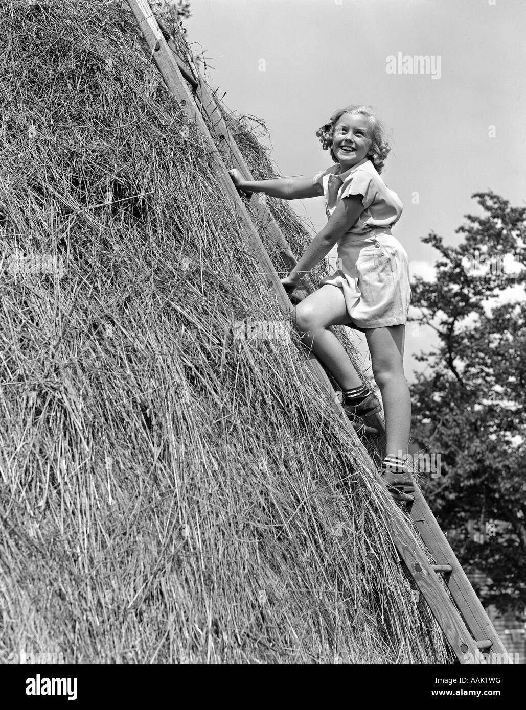 1940s sorridente ragazza bionda salendo la scala appoggiata contro il fieno di alta pila Foto Stock