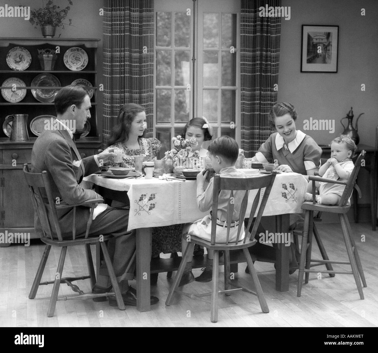1930s famiglia di 6 seduta al tavolo IN UNA SALA DA PRANZO DI MANGIARE LA PRIMA COLAZIONE il bambino è seduto in una sedia alta Foto Stock