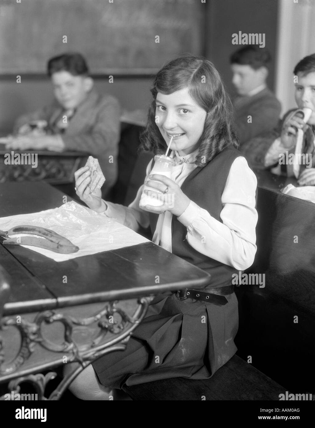 1920s School girl mangiare il pranzo alla sua scrivania di bere da una bottiglia di latte tenendo un sandwich Foto Stock