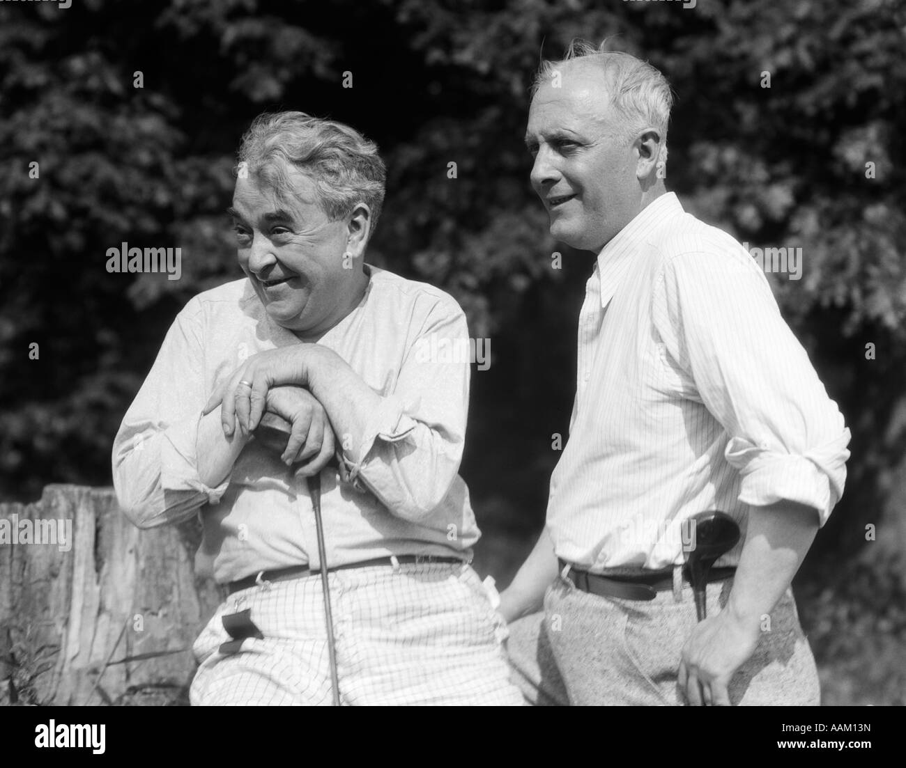 1920s 1930 2 uomini anziani holding club di golf seduti su un ceppo di albero GUARDANDO VERSO IL CAMPO DA GOLF Foto Stock