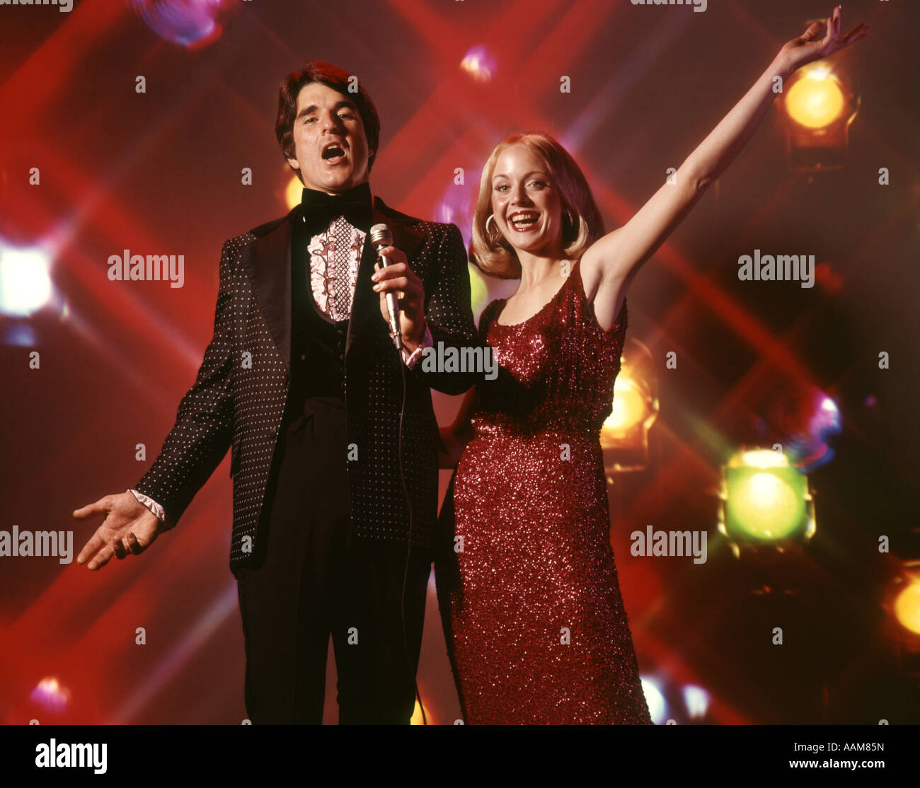 1970 anni settanta DUO uomo donna giovane cantando donna abito rosso abito uomo TUXEDO tenendo il microfono stadio sullo sfondo delle luci Foto Stock