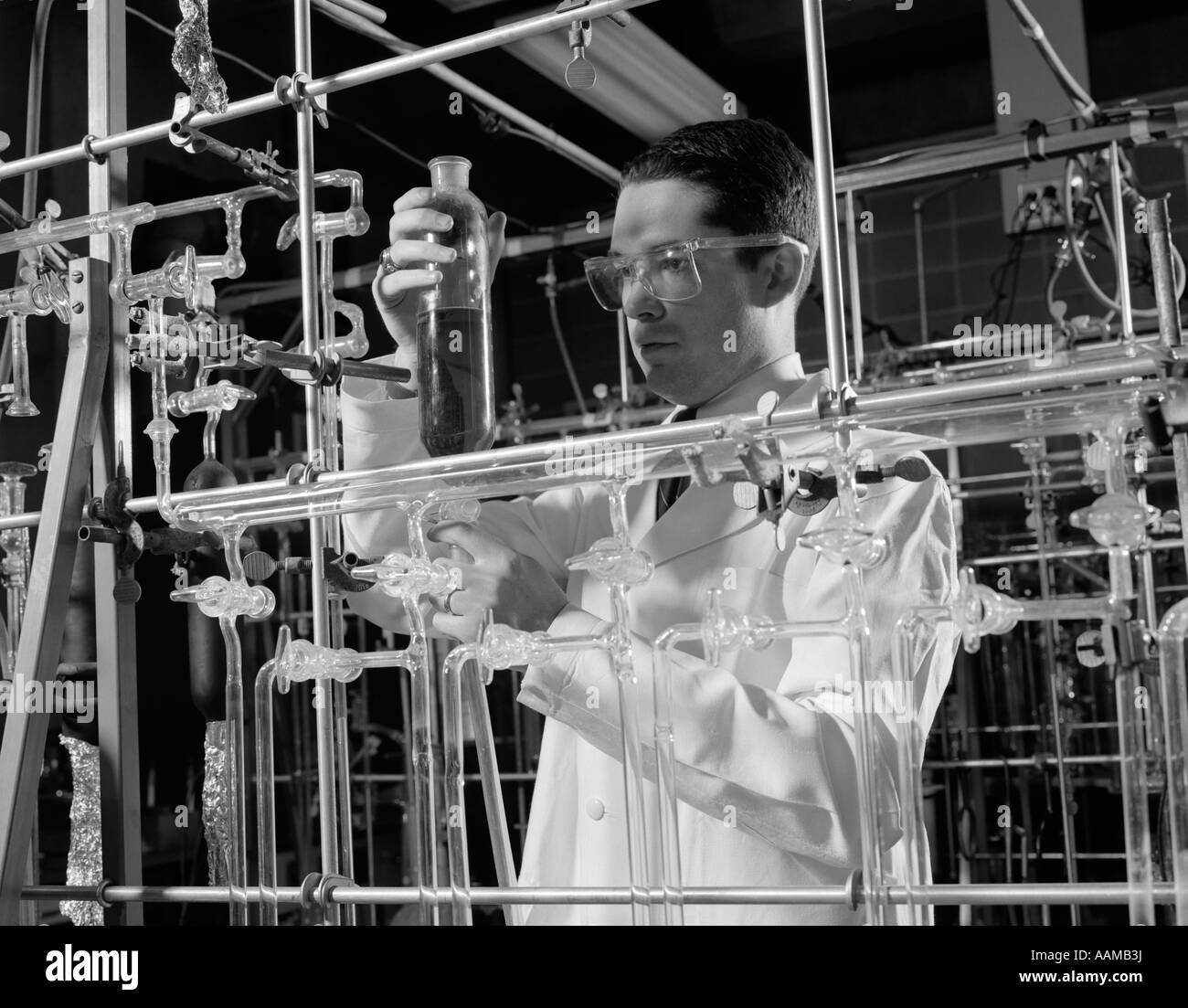 Anni sessanta uomo in camice da laboratorio con gli occhiali e circondato da tubi di vetro di condurre una ricerca scientifica Foto Stock