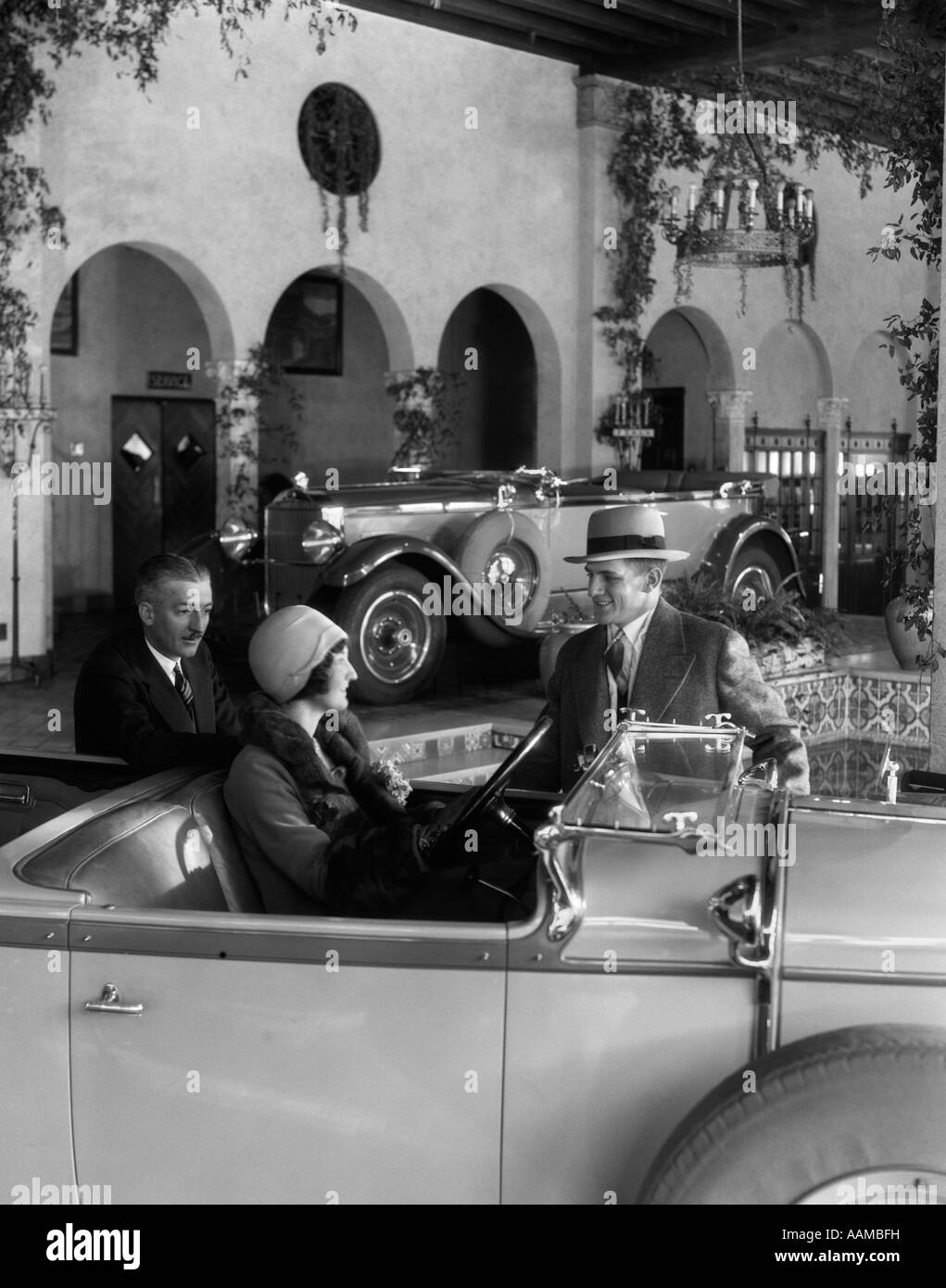 1920s DONNA NEL SEDILE DEL CONDUCENTE DI CONVERTIBLE con marito e venditore in piedi accanto a AUTO IN SHOWROOM DI FANTASIA Foto Stock