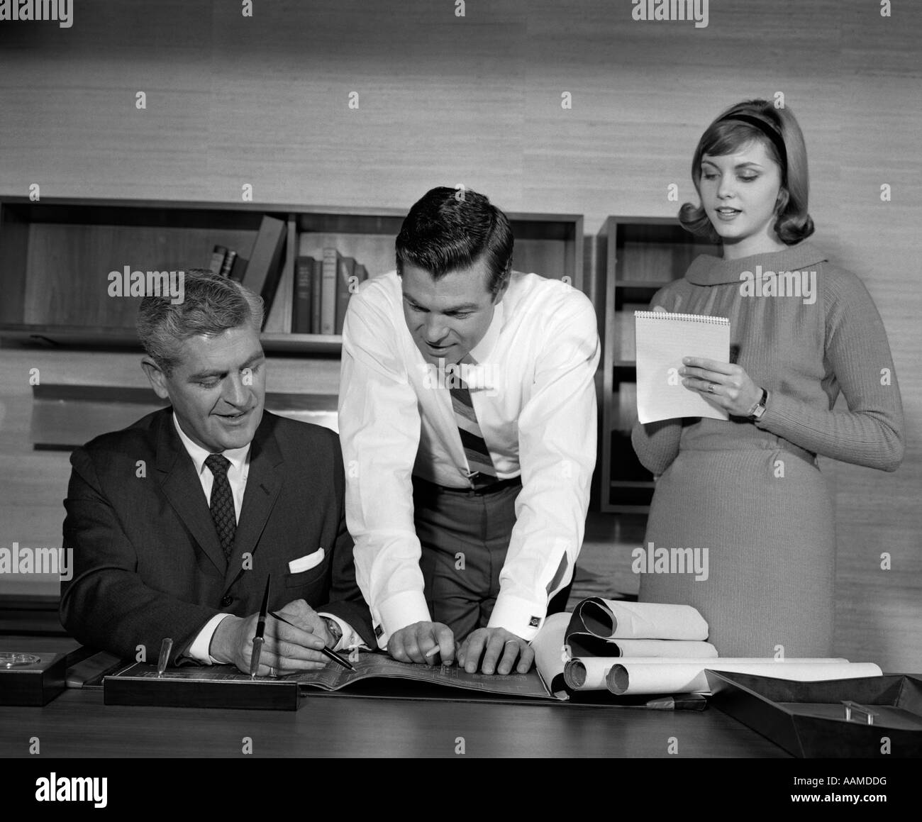 Anni sessanta COPPIA DI UOMINI DI AFFARI dietro la scrivania guardando Blueprint con il segretario a lato prendendo appunti Foto Stock