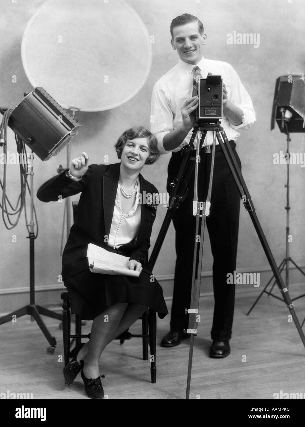 1920 Fotografo in piedi dietro la telecamera in studio con assistente seduto accanto a lui tenendo la documentazione Foto Stock