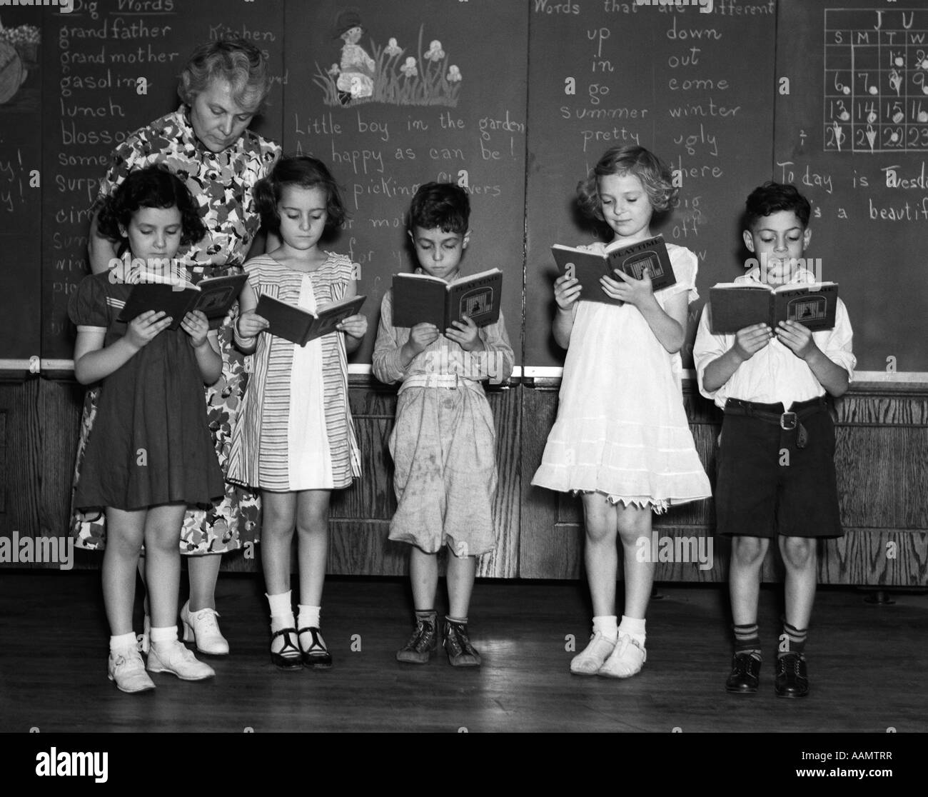 1930s line-up di 5 GLI STUDENTI DELLA SCUOLA ELEMENTARE DI FRONTE BLACKBOARD la lettura di libri con insegnante cercando su Foto Stock