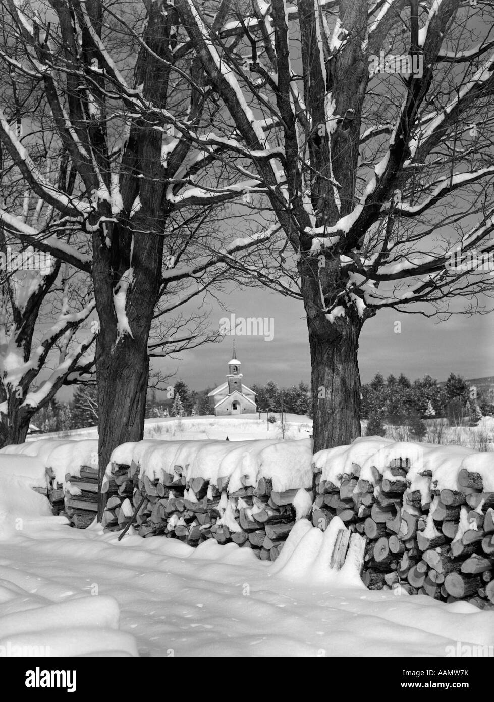 1940s pila di coperte di neve di legna da ardere LOGS impilati tra due alberi CON LA CHIESA DI CAMPAGNA IN BACKGROUND Foto Stock